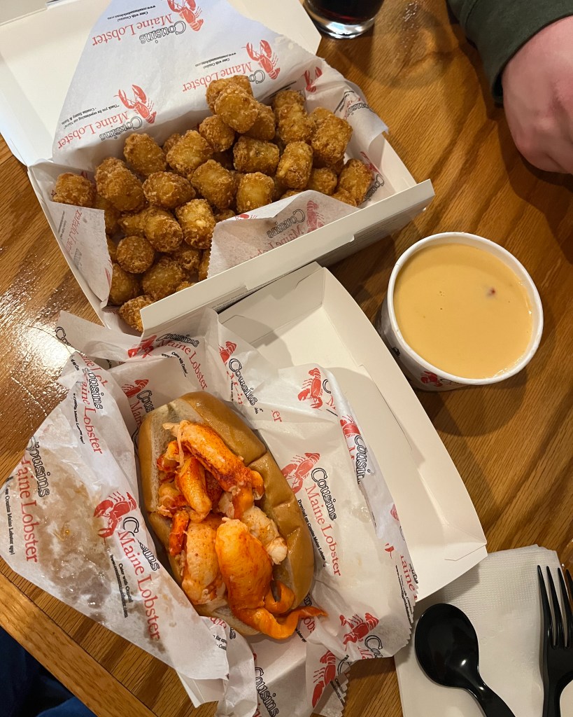 a lobster roll, cup of lobster bisque, and container of tater tots are spread out on a bar table